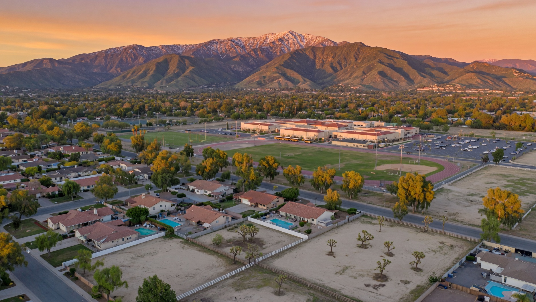 Aerial view of Quartz Hill residential neighborhood at sunset with snow-capped San Gabriel Mountains in background, showing single-family homes with pools, tree-lined streets, sports facilities, and commercial buildings